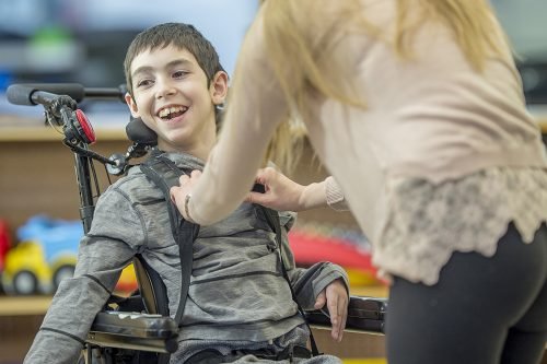 A special needs boy is in his classroom with an assistant. She is holding his hand, and he is smiling.